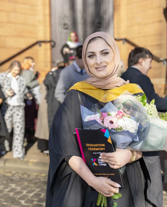 Female graduate wearing a hijab and gown, holding flowers and a certificate outside the graduation venue surrounded by well-wishers. Female graduate wearing a hijab and gown, holding flowers and a certificate outside the graduation venue surrounded by well-wishers.